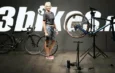 Une femme tient une roue de vélo au milieu d'un atelier avec deux vélos, dont un sur un pied de réparation.