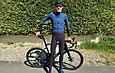 Cycliste debout avec un vélo noir devant une haie verte, portant une tenue Q36.5 bleue et noire.