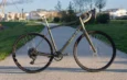 Vélo gravel vert olive avec selle et guidoline beiges, stationné de profil sur une allée goudronnée.