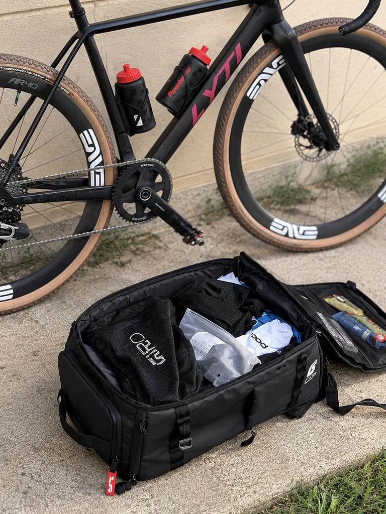 Scicon Rain Pro 35L ouvert et chargé avec des vêtements et accessoires de cyclisme à côté d’un vélo gravel. Exemple réel de son utilisation pour se rendre en compétition.