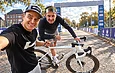 Deux hommes en selfie, l'un pointe un vélo gravel blanc sur piste caillouteuse, arche en fond.