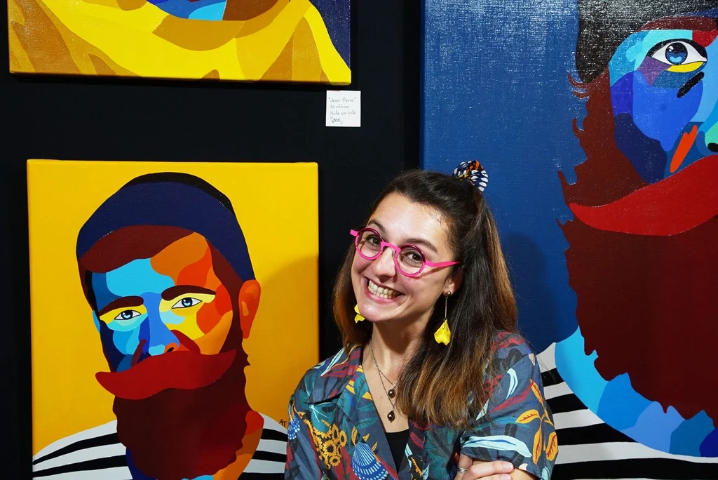 Mathilde AT pose devant plusieurs de ses peintures de portraits aux couleurs vives, dans un espace d'exposition. Un aperçu de son univers artistique.