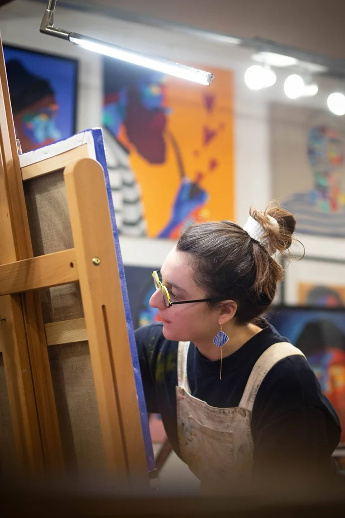Mathilde AT en plein travail artistique devant une toile installée sur un chevalet. Dans son atelier, entourée de peintures colorées, l’artiste rochelaise donne forme à ses créations.