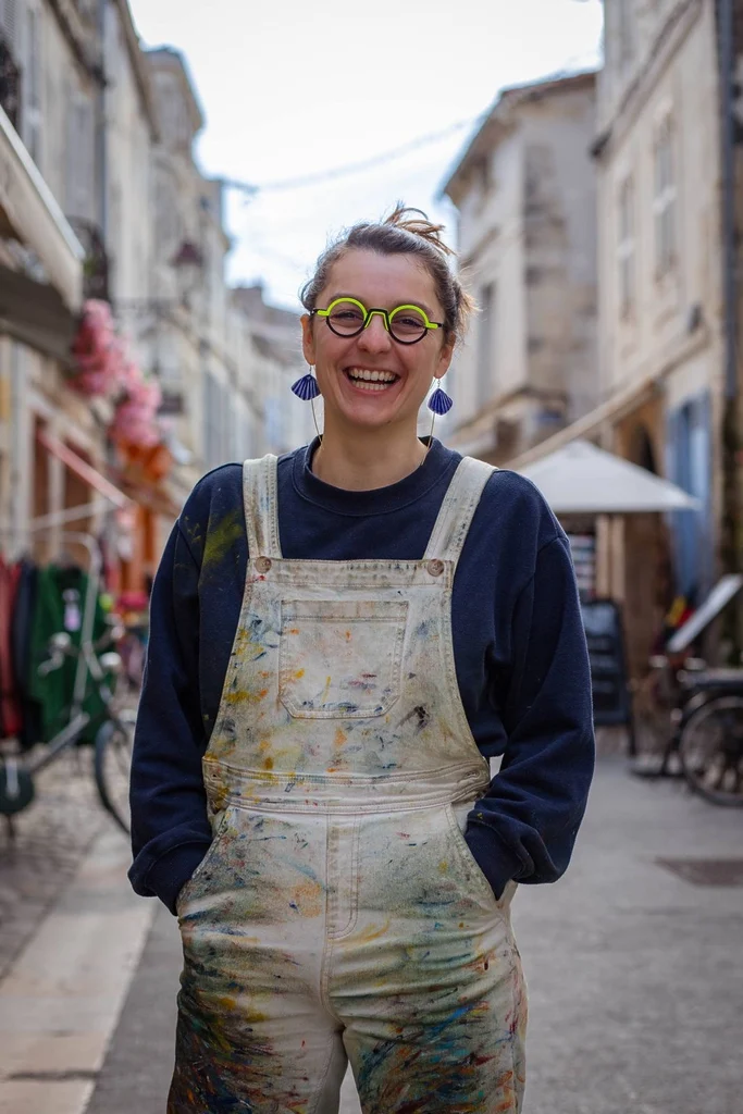 Portrait de Mathilde AT souriante dans une rue de La Rochelle, vêtue d’une salopette tachée de peinture, avec des lunettes vertes et des boucles d’oreilles bleues. Une photographie qui illustre l’univers coloré et la personnalité de l’artiste peintre.