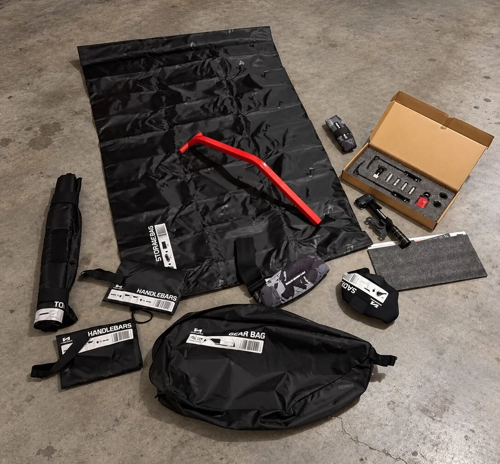 Ensemble des accessoires livrés avec la housse Scicon Aerocomfort 3.1 Gravel, disposés sur le sol en béton d'un garage : protections de cadre et de guidon, sangles, adaptateurs d'axes, micro-pompe, sac de rangement et accessoires de transport.