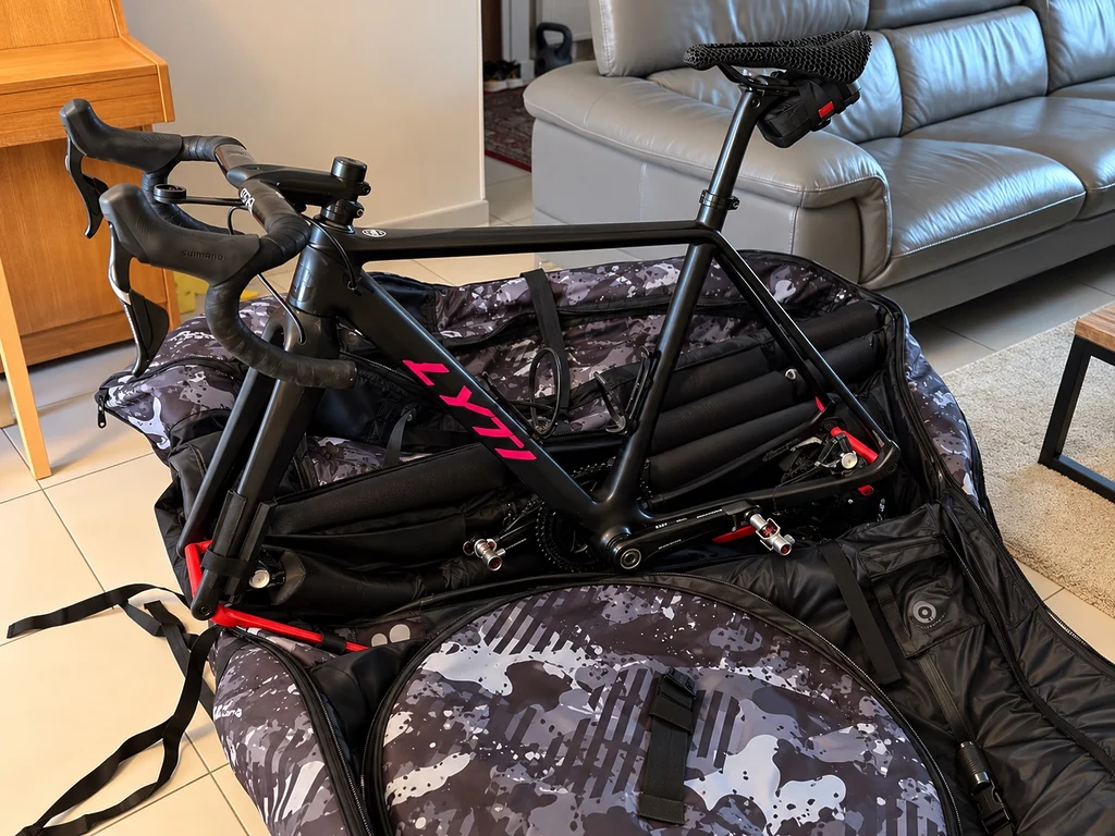 Gravel Lyti noir avec marquage rose installé sans ses roues dans la housse Scicon Aerocomfort 3.1 Gravel Grey Camo ouverte, montrant le cadre, le poste de pilotage et les éléments de maintien.