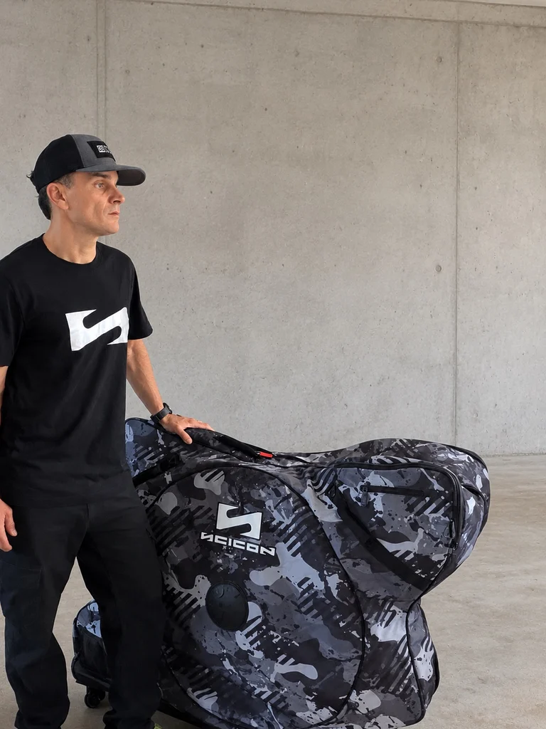 Jeff Tatard debout à côté de la housse de transport Scicon Aerocomfort 3.1 Gravel Grey Camo, une main posée sur le bagage, portant un tee-shirt et une casquette Scicon, devant un mur en béton gris.