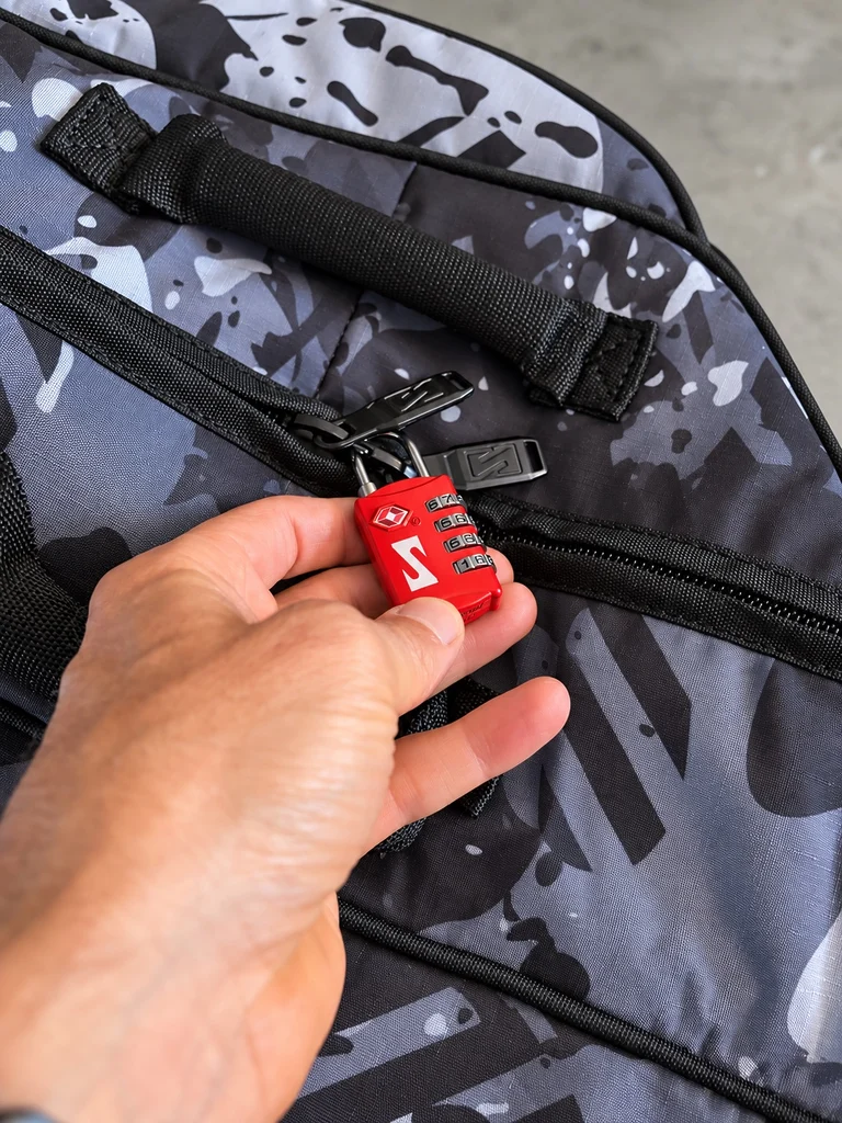 Cadenas rouge TSA Scicon à combinaison de quatre chiffres, tenu à la main devant la fermeture TwistZip de la housse Aerocomfort 3.1 Gravel en finition Grey Camo.