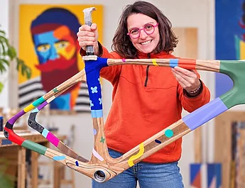Une femme aux lunettes roses pose avec un cadre de vélo en bois peint et coloré dans un atelier.